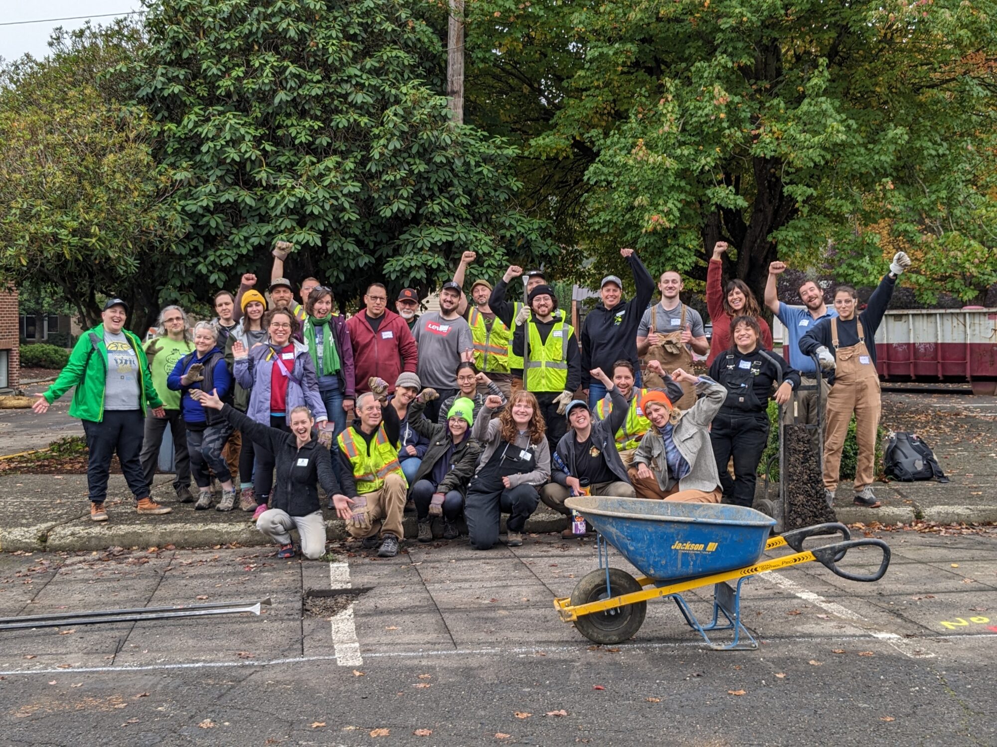 Group of 30 volunteers posing for a group photos. Volunteers are smiling with hands raised in the air. In front of the volunteers are tools used for removing concrete and a wheel barrow. 