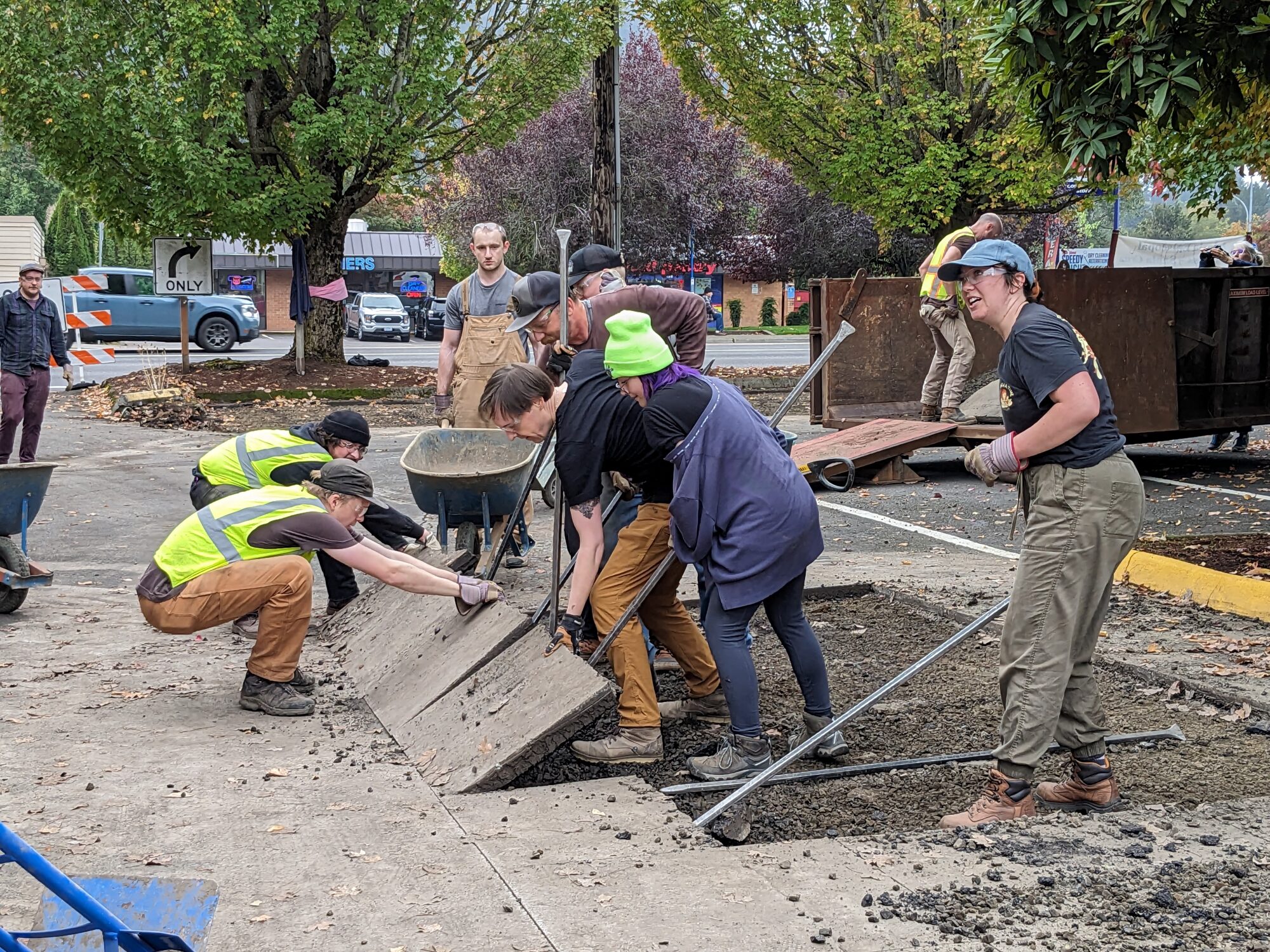 A group of 8 volunteers work together to remove a large piece of concrete. 2 volunteers are on the left side of the concrete pulling towards them while 4 volunteers on hte rights side of the slab of conrete use pry bars to push. 