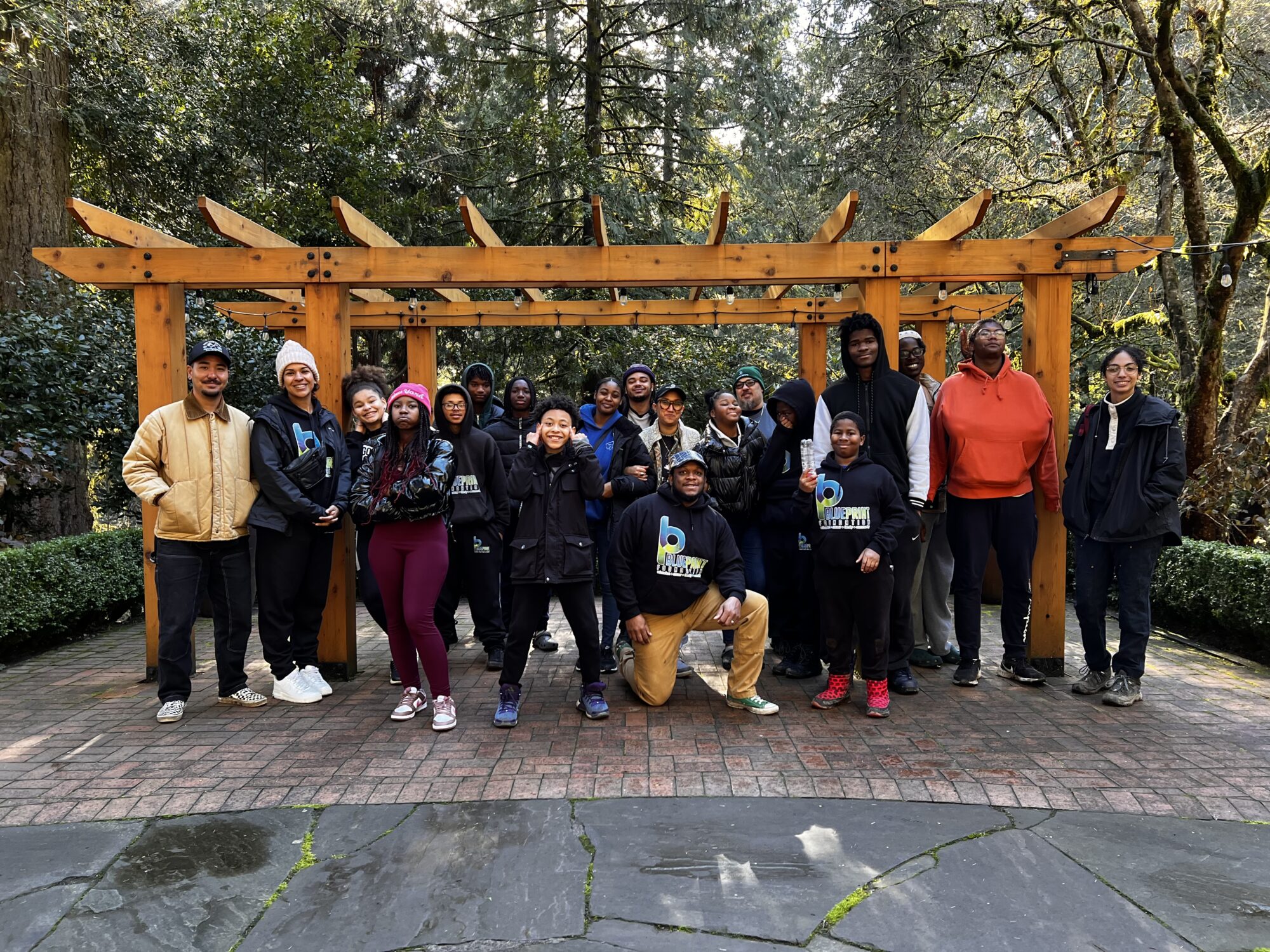 Group of 20 Blueprint Foundation youth and mentors. The group is posed in front of a pergola in a courtyard at Leach Botanical Garden with vegetation visible in the background. 