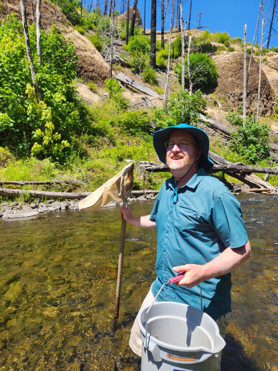 Photo of the author standing in a stream with a net and bucket for catching aquatic insects.